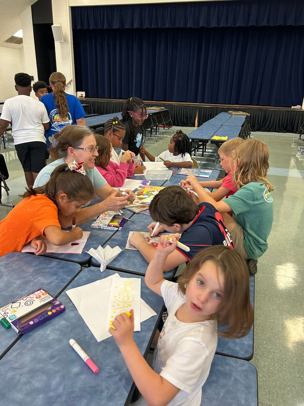 Kids coloring with markers at a long table while a staff member helps